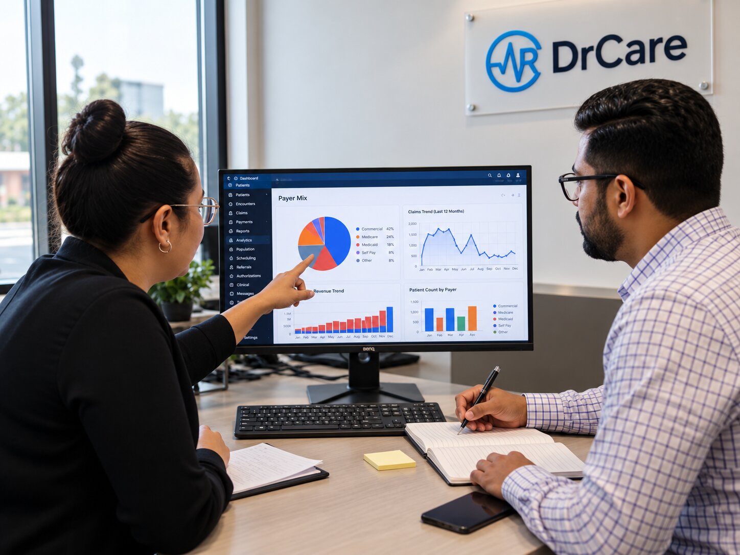 Two billing analysts discussing payer-mix data and trend charts on a monitor in a healthcare office